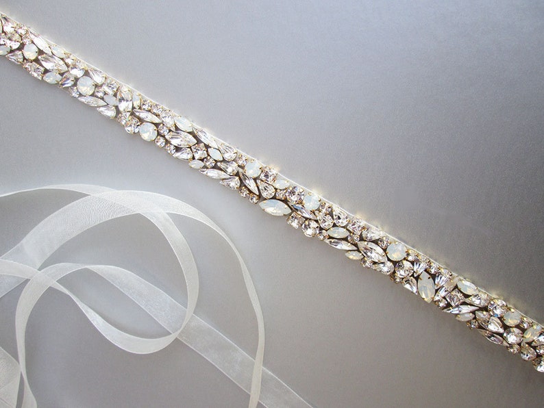 May include: A white satin ribbon with a gold and crystal embellishment. The embellishment is made up of small, clear crystals and gold beads.