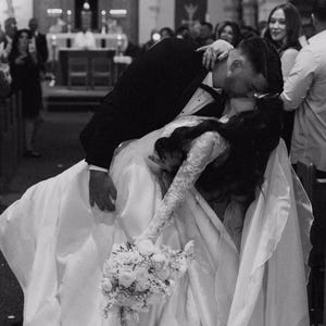 Pu&ograve; includere: Una sposa e uno sposo si baciano durante la loro cerimonia di matrimonio in una chiesa. La sposa indossa un abito da sposa bianco con un lungo strascico e un bouquet di fiori bianchi. Lo sposo indossa un abito nero e una camicia bianca. La coppia &egrave; circondata da ospiti.
