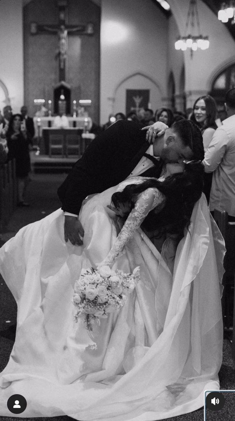 Pu&ograve; includere: Una sposa e uno sposo si baciano durante la loro cerimonia di matrimonio in una chiesa. La sposa indossa un abito da sposa bianco con un lungo strascico e un bouquet di fiori bianchi. Lo sposo indossa un abito nero e una camicia bianca. La coppia &egrave; circondata da ospiti.