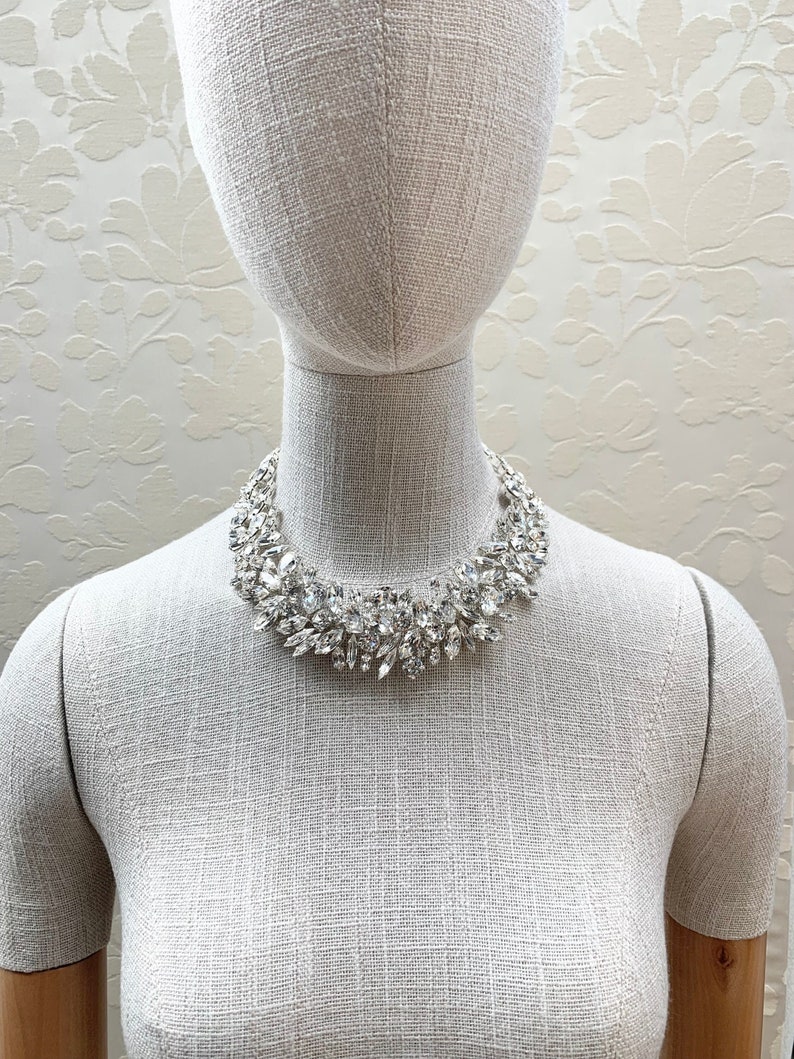 May include: A silver statement necklace with a cluster of clear and pearlescent crystals. The necklace is on a white mannequin.