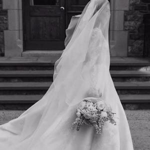 Pu&ograve; includere: Una sposa in abito da sposa bianco e velo tiene un bouquet di fiori bianchi. &Egrave; in piedi su una scalinata davanti a una porta.