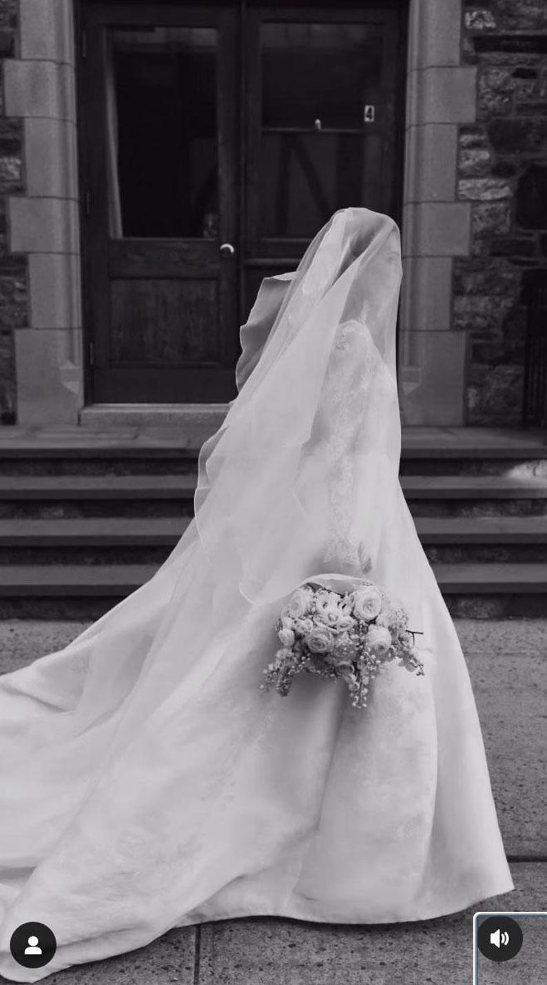 Pu&ograve; includere: Una sposa in abito da sposa bianco e velo tiene un bouquet di fiori bianchi. &Egrave; in piedi su una scalinata davanti a una porta.