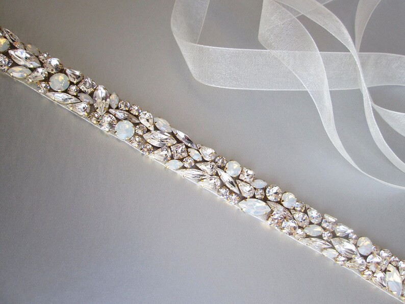 May include: A white satin ribbon with a row of clear and iridescent crystals sewn on top. The ribbon is on a gray background.