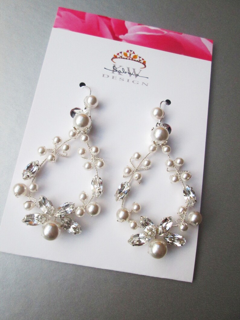 May include: A pair of silver earrings with a delicate floral design featuring small white pearls and clear crystals. The earrings are displayed on a white card with the text "KW DESIGN" printed on it.