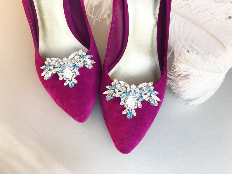 May include: A pair of fuchsia-colored high heel shoes with a jeweled embellishment on each toe. The embellishments are made of clear and blue rhinestones.