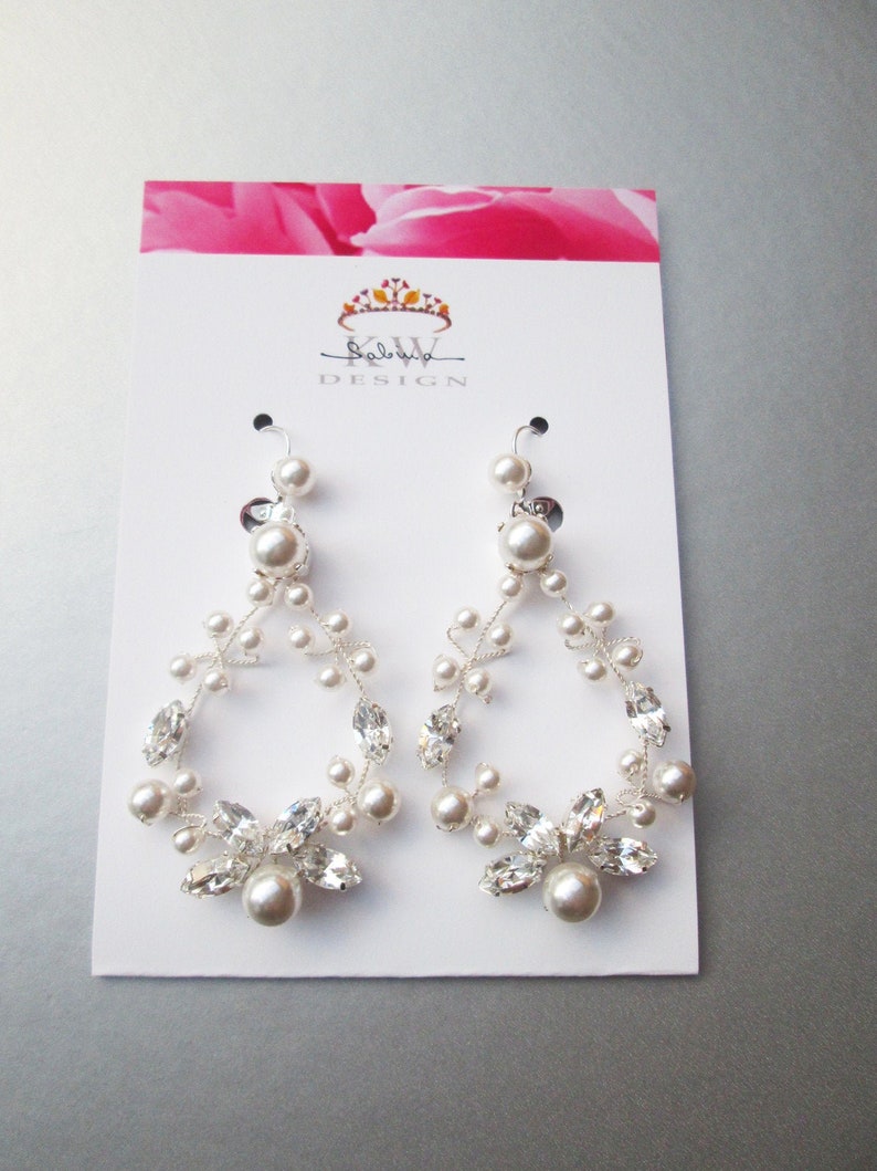 May include: A pair of silver earrings with a teardrop shape. The earrings are adorned with small white pearls and clear crystals. The earrings are displayed on a white card with the text "KW DESIGN" printed on it.