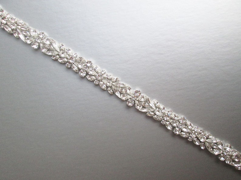 May include: A silver rhinestone trim with a floral design. The trim is made of small, clear rhinestones that are arranged in a delicate pattern.