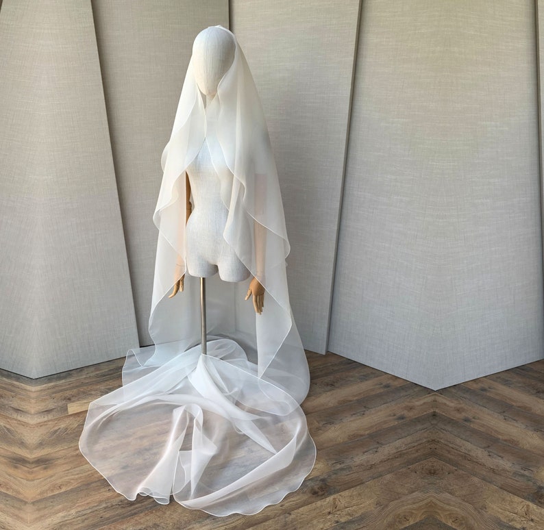 Pu&ograve; includere: Un velo da sposa in tulle bianco con un lungo strascico fluido. Il velo &egrave; drappeggiato su un manichino ed &egrave; esposto su uno sfondo grigio chiaro.
