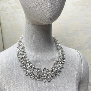 May include: A silver necklace with a large, statement design featuring clear, faceted crystals. The necklace is displayed on a white mannequin.