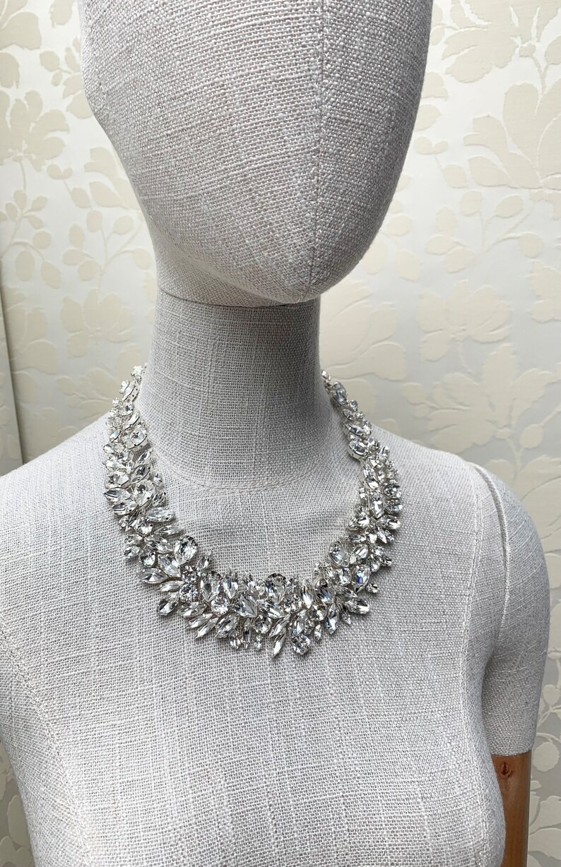 May include: A silver necklace with a large, statement design featuring clear, faceted crystals. The necklace is displayed on a white mannequin.
