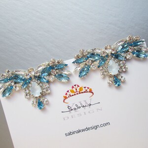 May include: A pair of silver earrings with blue and clear crystal embellishments. The earrings are shaped like a cluster of flowers with a large, pear-shaped crystal in the center of each cluster. The earrings are on a white card with the text "Sabina KW DESIGN" and "sabinakwdesign.com".