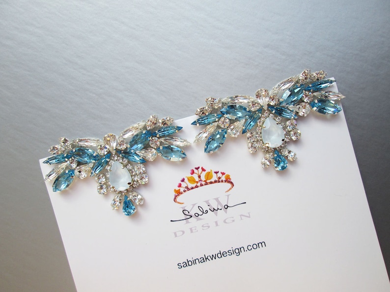 May include: A pair of silver earrings with blue and clear crystal embellishments. The earrings are shaped like a cluster of flowers with a large, pear-shaped crystal in the center of each cluster. The earrings are on a white card with the text "Sabina KW DESIGN" and "sabinakwdesign.com".