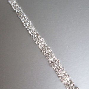 May include: A silver rhinestone trim with a floral design. The trim is made of small, clear rhinestones that are arranged in a repeating pattern of leaves and flowers.