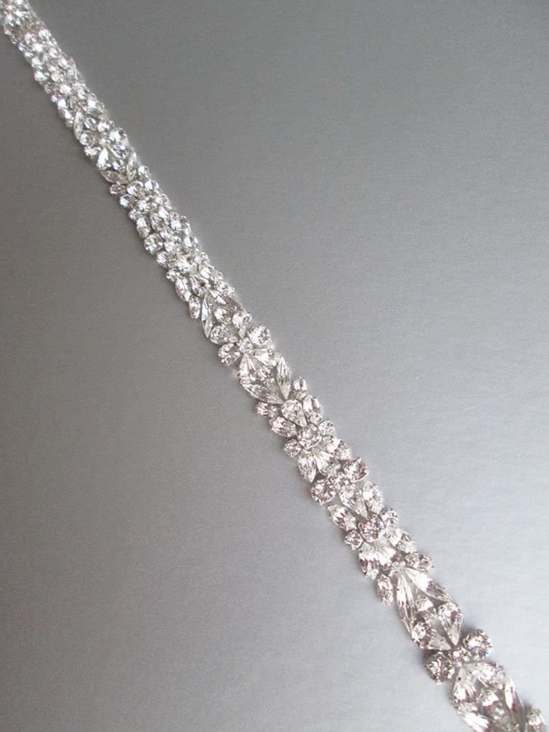 May include: A silver rhinestone trim with a floral design. The trim is made of small, clear rhinestones that are arranged in a repeating pattern of leaves and flowers.