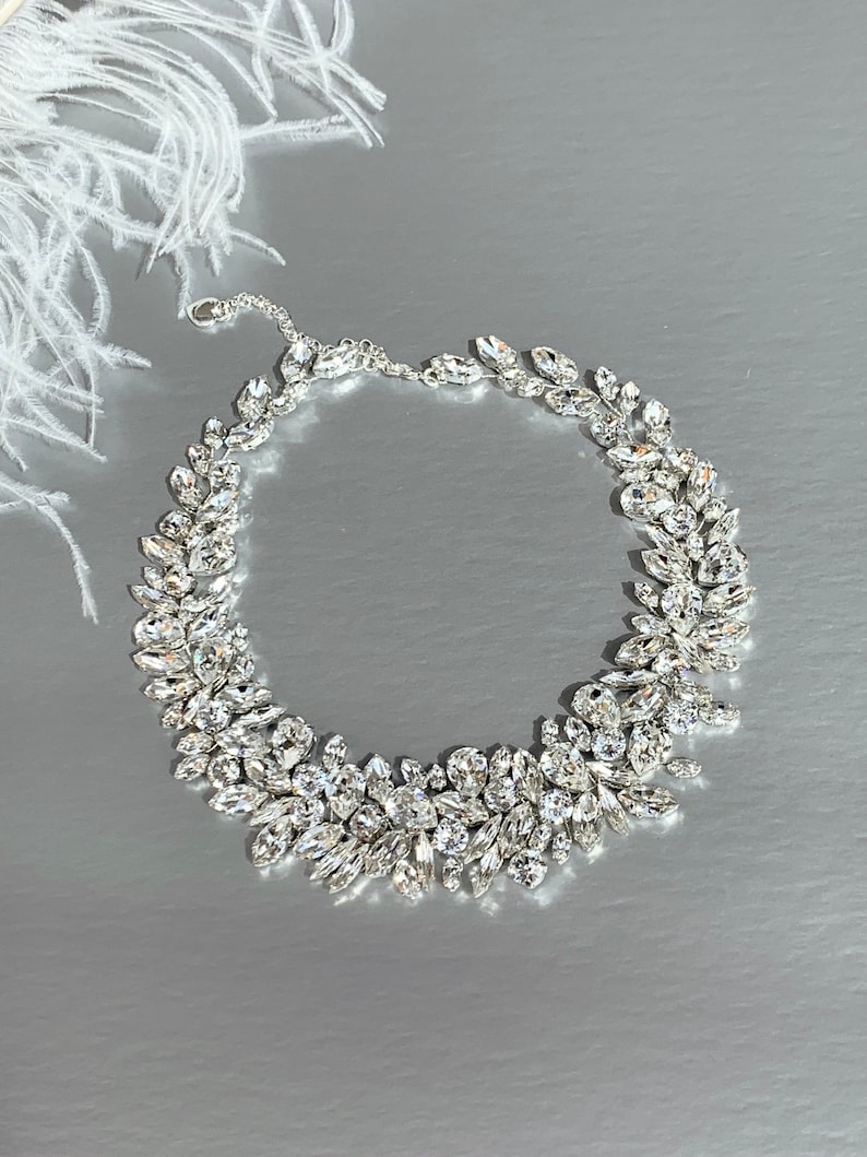 May include: A silver necklace with a delicate chain and a large, ornate design of clear crystal stones. The stones are arranged in a cluster of leaves, creating a flowing, organic look.