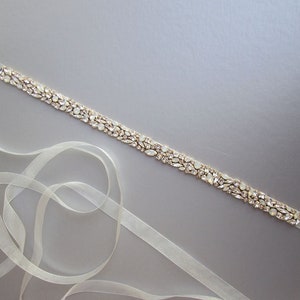 May include: A delicate ivory satin ribbon with a gold-toned embellishment of clear and iridescent rhinestones. The ribbon is approximately 1.5 inches wide and is perfect for adding a touch of sparkle to any outfit.