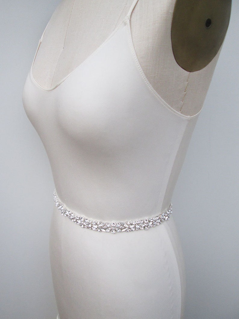 May include: A white satin bridal sash with a crystal embellishment. The sash is shown on a white dress form.