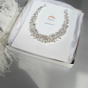 May include: A white box with a white tissue paper lining. Inside the box is a silver crystal necklace with a leaf design. The necklace is displayed on a white card with the text "Sabina Design" and a website address.