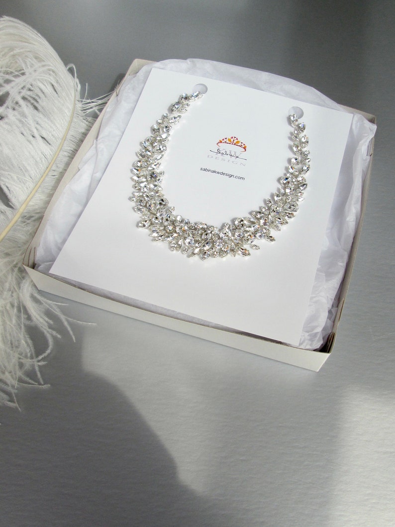 May include: A white box with a white tissue paper lining. Inside the box is a silver crystal necklace with a leaf design. The necklace is displayed on a white card with the text "Sabina Design" and a website address.