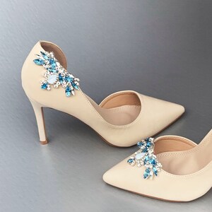 May include: A pair of beige high-heeled shoes with a pointed toe. Each shoe has a decorative cluster of blue and clear rhinestones on the top.