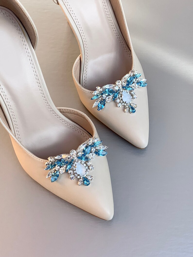 May include: A pair of beige high-heeled shoes with a blue and silver crystal embellishment on the toe.