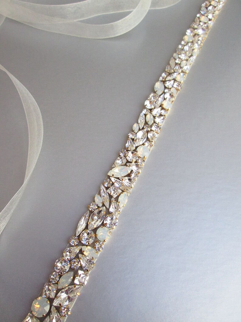 May include: A white satin ribbon embellished with a row of clear and iridescent rhinestones. The rhinestones are set in gold metal.