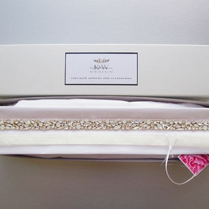 May include: A white box with a pink interior containing a delicate, crystal-encrusted bridal sash. The box has a label that reads "Bridal Design Fine Hair Jewelry and Accessories".
