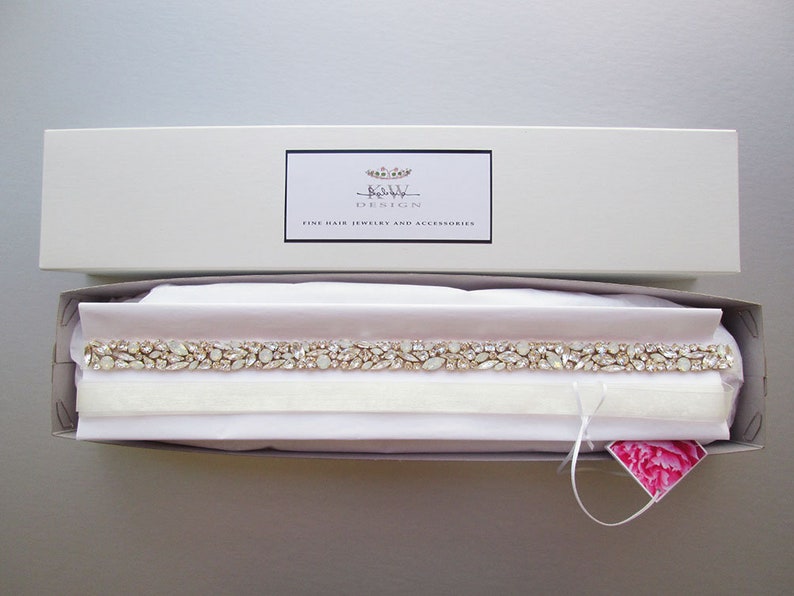 May include: A white box with a pink interior containing a delicate, crystal-encrusted bridal sash. The box has a label that reads "Bridal Design Fine Hair Jewelry and Accessories".
