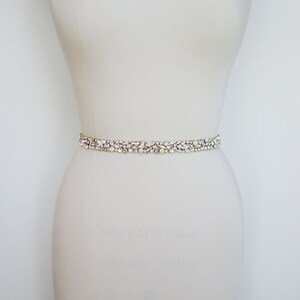 May include: A white bridal sash with a delicate, crystal-encrusted design. The sash is made of a thin, flexible material and is adorned with small, clear crystals that sparkle in the light.