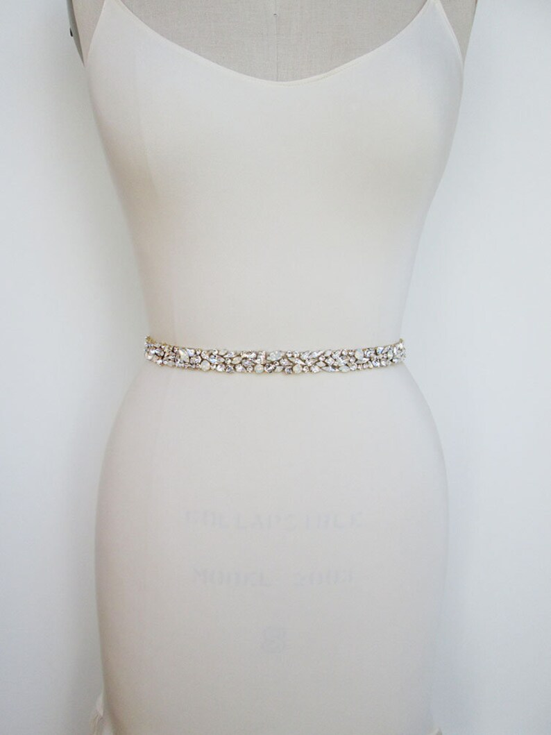May include: A white bridal sash with a delicate, crystal-encrusted design. The sash is made of a thin, flexible material and is adorned with small, clear crystals that sparkle in the light.