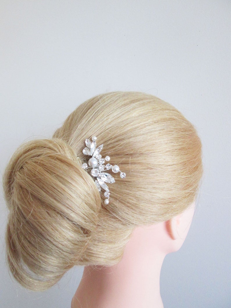 Puede incluir: Un peine de pelo plateado con cristales y perlas transparentes y blancas. El peine tiene forma de flor con una base puntiaguda.