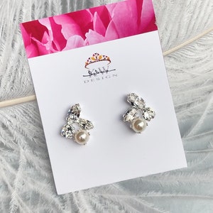 May include: A pair of silver earrings featuring a cluster of clear rhinestones and a single pearl at the centre. The earrings are presented on a white card with the text "Salvia Design".