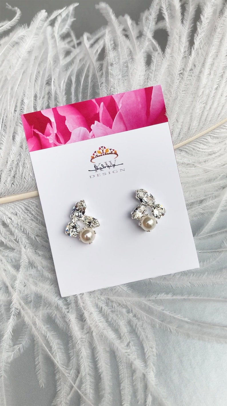 May include: A pair of silver earrings featuring a cluster of clear rhinestones and a single pearl at the centre. The earrings are presented on a white card with the text "Salvia Design".