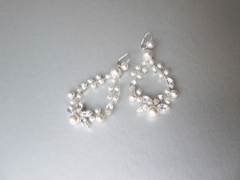 May include: A pair of silver earrings with a delicate design featuring white pearls and clear crystals. The earrings are shaped like a teardrop and dangle from the earlobe.