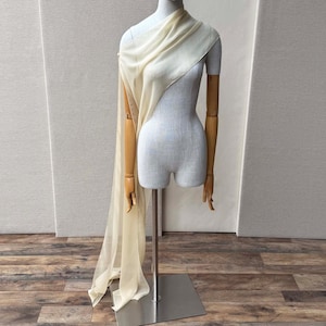 May include: A cream-colored, sheer fabric draped over a mannequin. The fabric cascades down the front, creating a flowing effect. The mannequin has wooden arms and is mounted on a metal stand. The background is a neutral color.