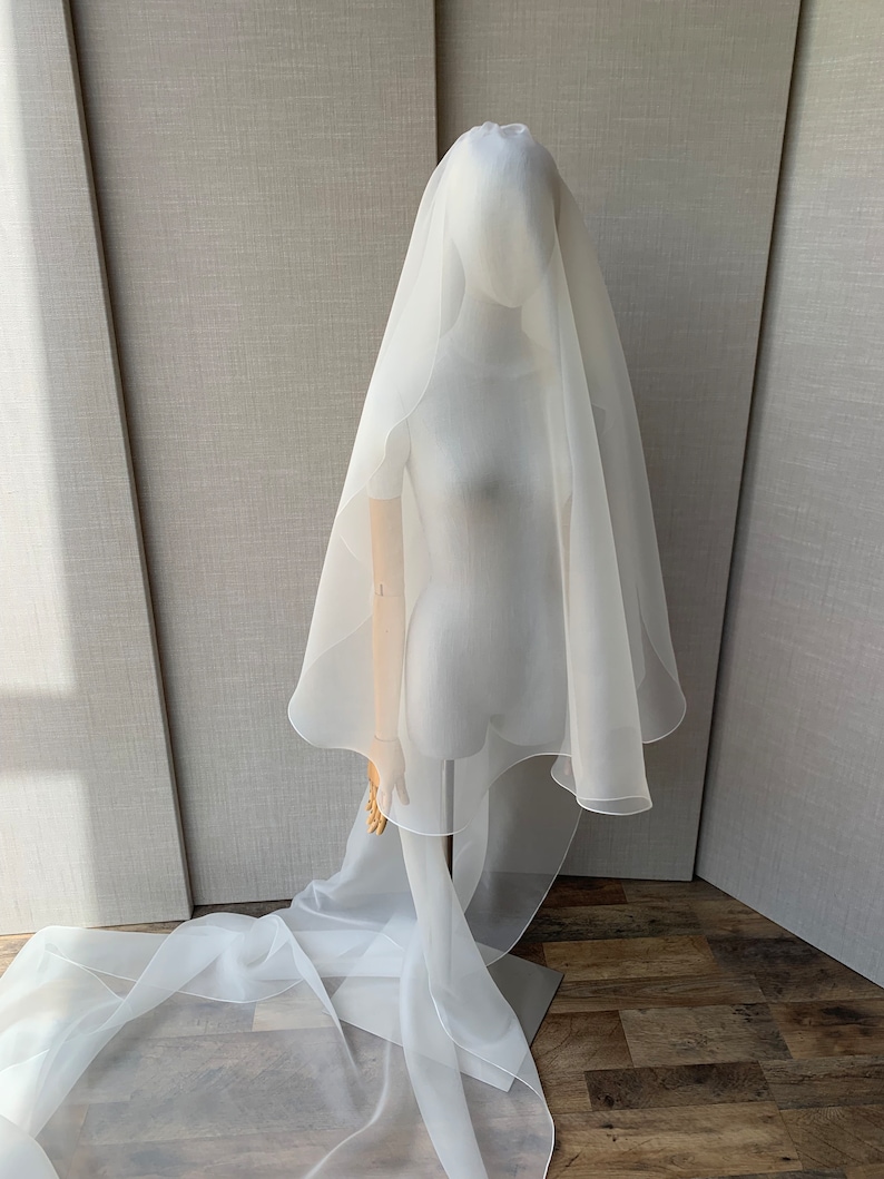 Pu&ograve; includere: Un velo da sposa in tulle bianco a strato singolo con bordo smerlato. Il velo &egrave; drappeggiato su un manichino e cade sul pavimento.