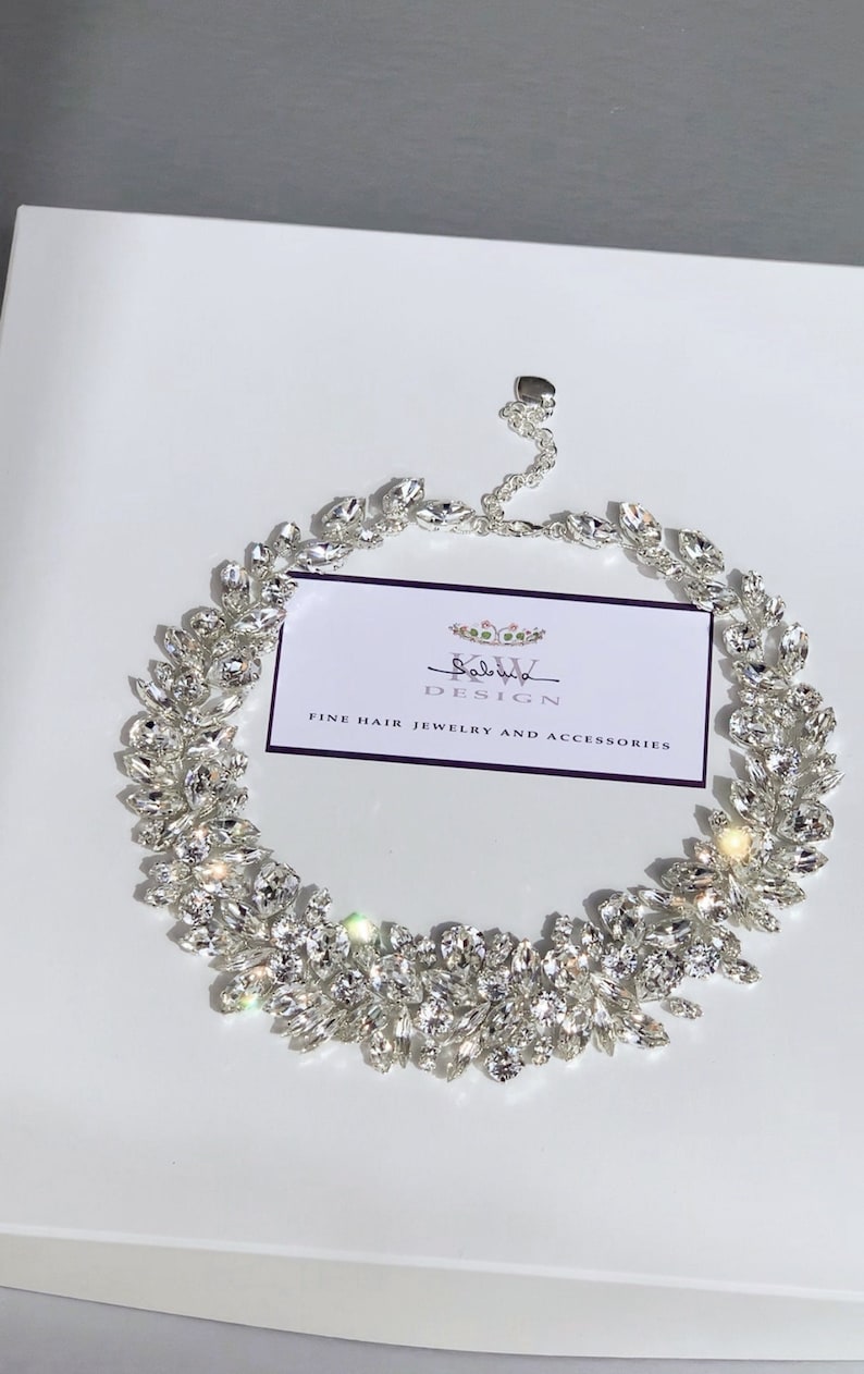May include: A silver necklace with a delicate chain and a large cluster of clear crystals in a leaf-like design. The necklace is displayed on a white background with a business card that reads "Salvatore Design Fine Hair Jewelry and Accessories".
