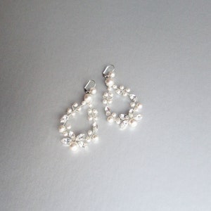 May include: A pair of silver earrings with a delicate design featuring small white pearls and clear rhinestones. The earrings are shaped like a teardrop and dangle from the ear.