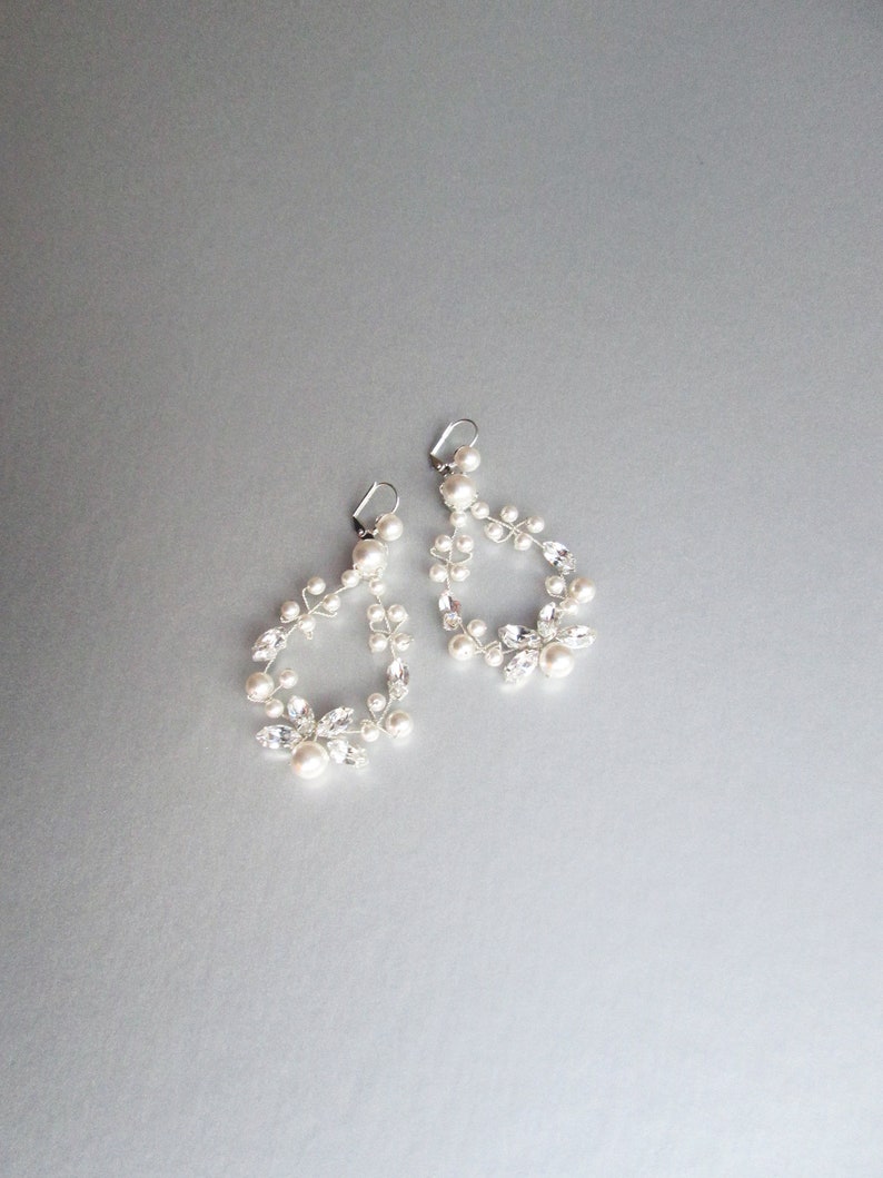 May include: A pair of silver earrings with a delicate design featuring small white pearls and clear rhinestones. The earrings are shaped like a teardrop and dangle from the ear.