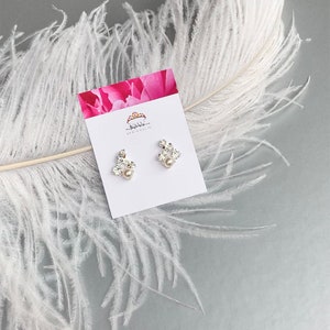 May include: A pair of silver earrings featuring a cluster of clear rhinestones and a single pearl at the centre. The earrings are presented on a white card with a pink floral design and the text "Shibboleth Jewelry".