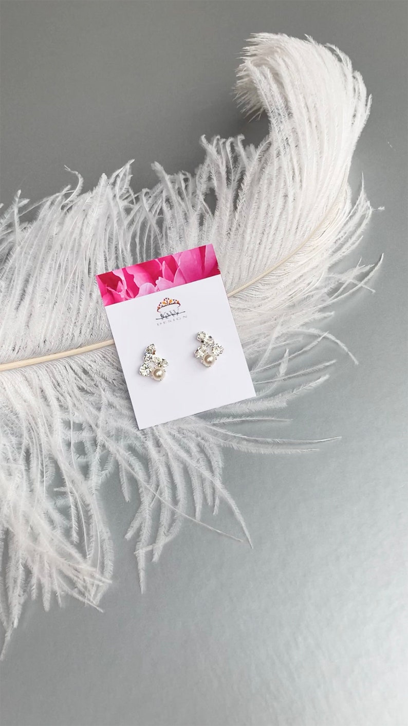 May include: A pair of silver earrings featuring a cluster of clear rhinestones and a single pearl at the centre. The earrings are presented on a white card with a pink floral design and the text "Shibboleth Jewelry".