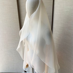 Pu&ograve; includere: Un velo da sposa in tulle bianco con un design a cascata a pi&ugrave; strati. Il velo &egrave; drappeggiato sulla testa e sulle spalle di un manichino, mostrando la sua texture delicata e fluida.