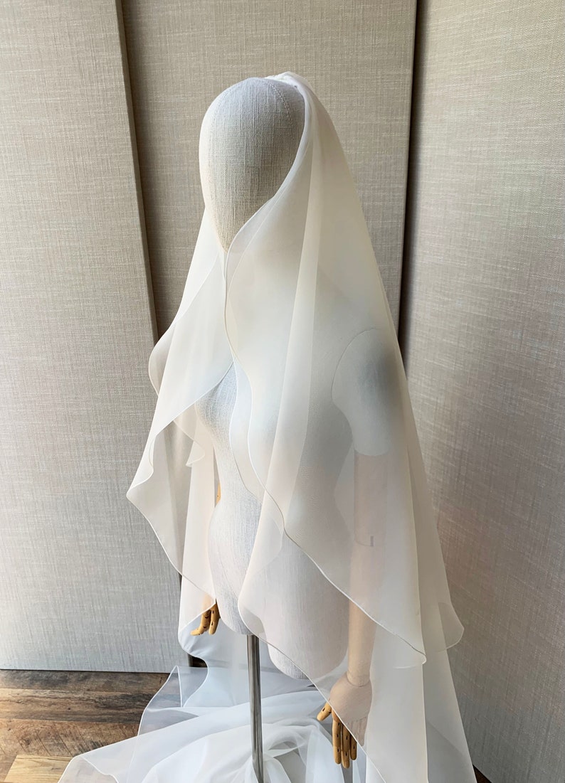 Pu&ograve; includere: Un velo da sposa in tulle bianco con un design a cascata a pi&ugrave; strati. Il velo &egrave; drappeggiato sulla testa e sulle spalle di un manichino, mostrando la sua texture delicata e fluida.