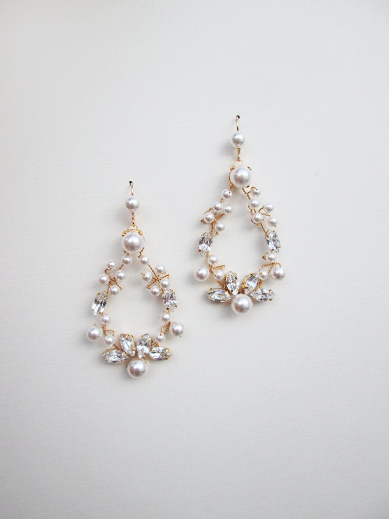 May include: A pair of gold dangle earrings with a teardrop shape. The earrings are adorned with small white pearls and clear rhinestones.