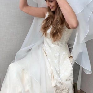 Pu&ograve; includere: Un abito da sposa bianco con un motivo in pizzo e un velo lungo e fluido. L'abito ha un corpetto aderente e una gonna ampia.