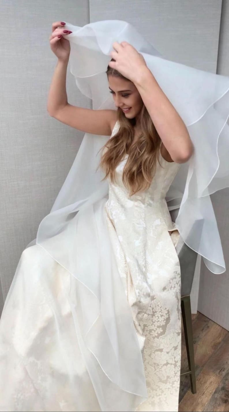 Pu&ograve; includere: Un abito da sposa bianco con un motivo in pizzo e un velo lungo e fluido. L'abito ha un corpetto aderente e una gonna ampia.