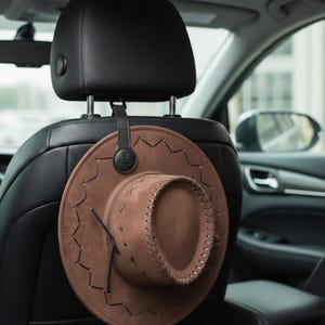 Puede incluir: Un sombrero de vaquero marrón cuelga de un reposacabezas negro de coche. El sombrero es de fieltro y tiene un ala y una corona cosidas. Una correa de cuero negro con un emblema de ancla asegura el sombrero.