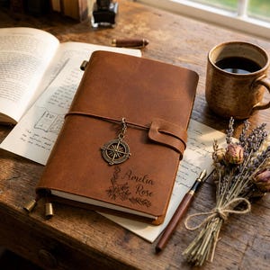 May include: A brown leather journal featuring a metal compass charm attached to a leather strap closure. The cover is engraved with the name Amelia Rose and a floral design. The journal is positioned on a desk with a fountain pen and book.