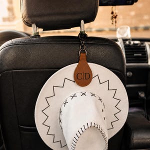 Può includere: Un cappello da cowboy bianco con cuciture nere e un'etichetta in pelle marrone con le iniziali "CD" è appeso a un poggiatesta per auto. Il cappello ha un bordo a zig-zag ed è sospeso da una clip nera.