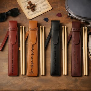 May include: Four leather drumstick cases in brown, tan, and black, each with a pair of drumsticks. The cases have personalized text, including names and quotes. Sunglasses, drum picks, and sheet music are also visible.